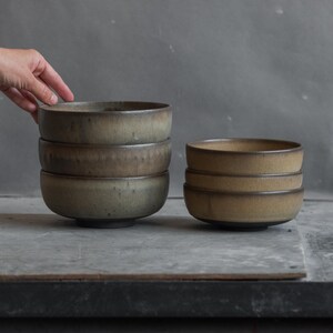 May include: A set of six ceramic bowls arranged in two stacks. The three larger bowls on the left feature a muted, earthy green and brown glaze with a dark rim. The three smaller bowls on the right have a warm, sandy tan finish with dark bases.