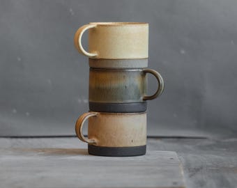 May include: Three ceramic mugs stacked on top of each other. The top mug is light beige with a dark brown rim. The middle mug is a gradient of brown, green, and beige with a dark brown rim. The bottom mug is light beige with a dark brown rim.