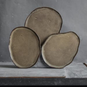 May include: Three ceramic plates in various sizes with an irregular, organic shape. The plates are a light brown color with a textured surface.