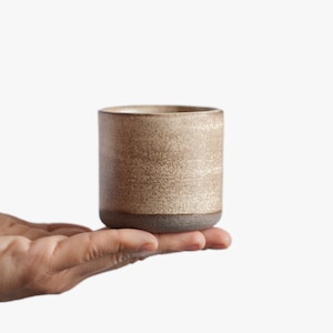 Op de afbeelding: Een klein, beige keramiek kopje met een bruine rand, vastgehouden in een hand.