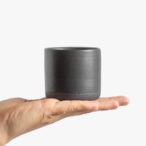 Op de afbeelding: Een donkergrijze keramische mok met een licht getextureerd oppervlak. De mok wordt in een hand gehouden.