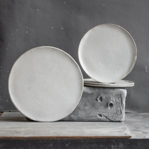 Könnte beinhalten: Drei weiße Keramikteller, die auf einem grauen Betonblock gestapelt sind. Die Teller sind rund und haben eine glatte, matte Oberfläche.