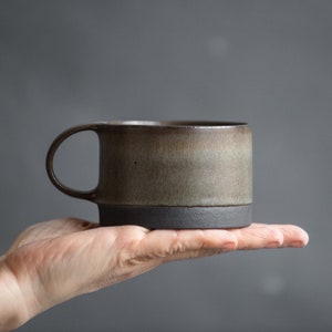 Puede incluir: Una taza de cerámica marrón y gris con un borde negro y un asa. La taza está en la mano de una persona.