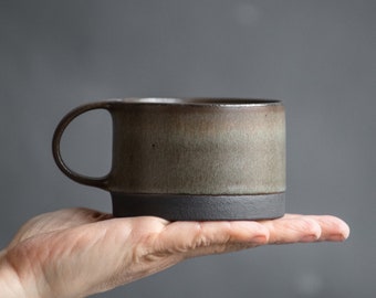 May include: A brown and gray ceramic mug with a black rim and a handle. The mug is sitting in a person's hand.