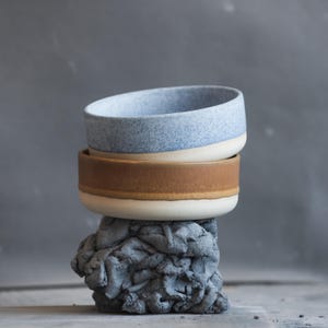 May include: Two ceramic bowls stacked on a gray, textured base. The top bowl is light blue with a speckled pattern, the middle bowl is brown, and both have a white rim. The base is a rough, gray sculpture.