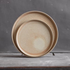 Può includere: Due piatti in ceramica, uno annidato nell'altro. I piatti sono di colore beige chiaro con una texture punteggiata.