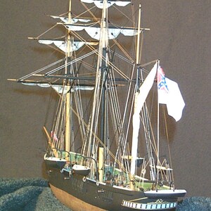 May include: A detailed model of a ship with white sails and a Confederate flag flying from the mast. The ship is black with a brown hull and is sitting on a dark grey surface.