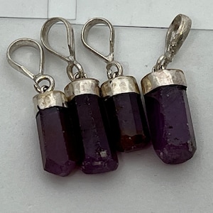 May include: Four purple gemstone pendants with silver metal loops. The gemstones are cylindrical with faceted sides. The silver loops are attached to the top of each gemstone. The pendants are displayed against a white background.