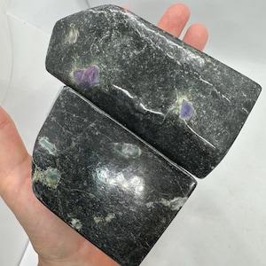 May include: Two dark green, rectangular stones with rounded edges. The stones have a speckled pattern with small patches of purple and green. The stones are held in a hand against a white background.