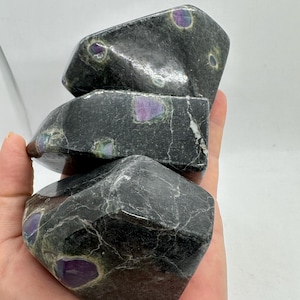 May include: Three dark grey, faceted stones with purple and green inclusions. The stones have a polished appearance and are stacked on top of each other. The stones are held in a hand against a white background.