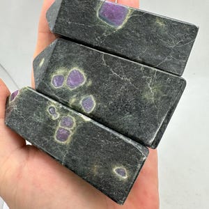 May include: Three rectangular, dark grey stone crystals with purple and white inclusions are held in a hand. The crystals have a polished finish and a slightly angled top. The stone has a marbled appearance.