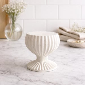 May include: A white, textured pedestal with a fluted design. The pedestal has a wide, flat top and a flared base. The product is likely used for displaying decorative items. The pedestal is made of a solid material.