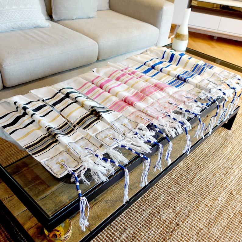 K&ouml;nnte beinhalten: Ein Satz von f&uuml;nf Tallit-Gebetsm&auml;nteln mit verschiedenen farbigen Streifen. Die Tallit sind auf einem Holztisch mit Glasplatte gestapelt. Die Tallit sind aus wei&szlig;em Stoff mit goldenen und blauen Streifen gefertigt. Die Tallit haben Fransen an den Enden.