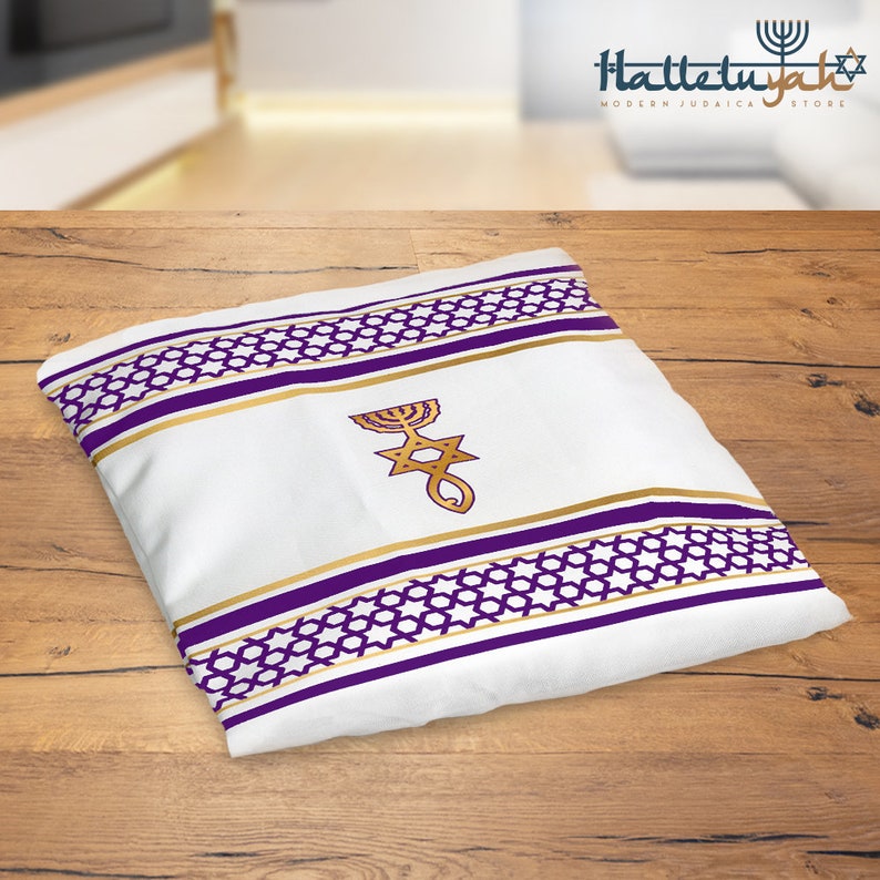 K&ouml;nnte beinhalten: Ein wei&szlig;er Tallit mit violetten und goldenen Streifen und einem Davidstern mit einem Menora-Design. Der Tallit liegt auf einer Holzoberfl&auml;che.