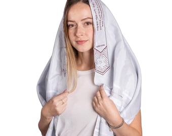 May include: A white prayer shawl with embroidered text and geometric designs in burgundy. The shawl is draped over the head and shoulders. The text on the shawl is in Hebrew. The person is wearing a white t-shirt.