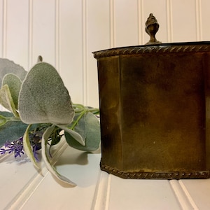 May include: A vintage brass container with a decorative finial on the lid. The container has an octagonal shape and a rope-like design around the base. A sprig of faux lavender and dusty miller is next to the container.