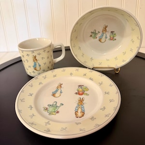 May include: A ceramic three-piece dinnerware set featuring a mug, a bowl, and a plate. Each piece is decorated with illustrations of a rabbit in a blue jacket, a green watering can, and potted plants against a pale yellow background with blue bows.