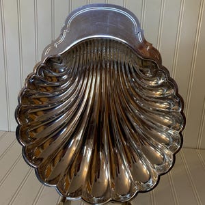 May include: A silver-colored, shell-shaped serving dish with a scalloped edge. The dish is supported by a decorative metal stand with curved legs. The interior of the dish has a ribbed design, reflecting light.