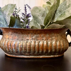 May include: An oval copper planter with a fluted body and a decorative etched band near the rim. The vessel features two dark metal ring handles on the sides and is filled with green artificial leaves and purple lavender sprigs.