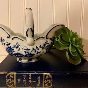 May include: A white porcelain gravy boat with a blue and white floral design. The boat has a handle and is sitting on a blue book. A green succulent plant is next to the boat.