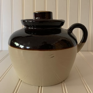 May include: A brown and white glazed ceramic crock with a lid and a handle. The crock is a traditional style with a rounded body and a wide mouth.