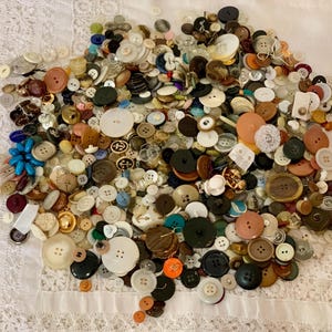 May include: A large assortment of vintage buttons in various sizes, shapes, and colors. The buttons include shades of brown, white, black, and blue, with some featuring decorative details. The buttons are scattered on a white lace fabric.