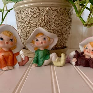 May include: Three ceramic elf figurines with painted details. The elf on the left is wearing a white hat and orange outfit. The elf in the middle is wearing a white hat and green outfit. The elf on the right is wearing a pink hat and brown outfit.
