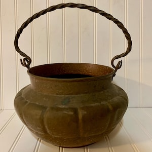 May include: A vintage bronze cauldron with a twisted metal handle. The cauldron has a rounded body with a textured surface and a dark interior. The handle is attached to the cauldron with decorative swirls.