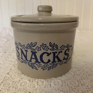 May include: A beige ceramic snack jar with a lid. The word "SNACKS" is printed in blue script, surrounded by a floral design. The jar is cylindrical and sits on a white lace surface.