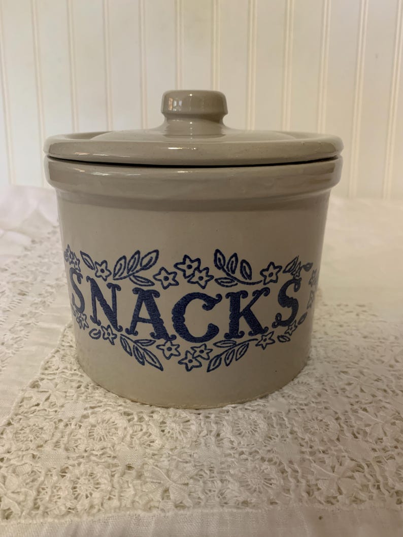 May include: A beige ceramic snack jar with a lid. The word "SNACKS" is printed in blue script, surrounded by a floral design. The jar is cylindrical and sits on a white lace surface.