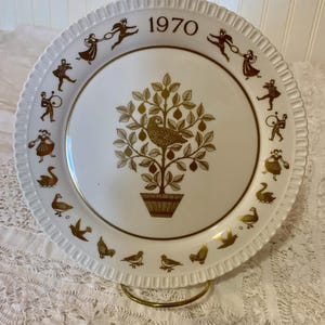 May include: A white porcelain plate with a gold rim and a gold design of a pear tree with a bird in the center. The plate has a gold stand and the year 1970 is written in gold at the top.