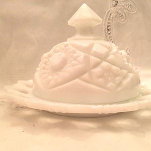 May include: A white milk glass butter dish featuring a dome-shaped lid with a geometric pressed pattern and a pointed finial. The base has a scalloped, openwork edge design. The entire piece is opaque white with a smooth, glossy finish throughout.