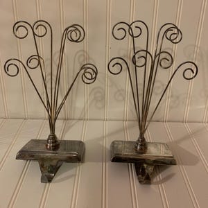 May include: A pair of silver stocking holders with a decorative swirl design. Each holder has a base with a hook for hanging stockings.