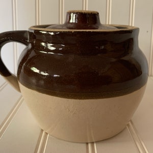 May include: A brown and white glazed ceramic crock pot with a lid. The crock pot has a rounded body and a single handle.