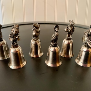 May include: Set of seven silver-toned bells, each topped with a detailed figurine of a different animal. The figurines include a rabbit, mouse, and duck. The bells are arranged on a dark surface.