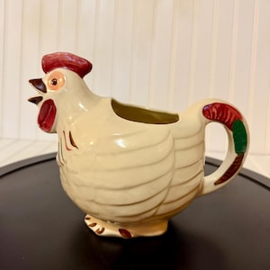 May include: A cream-colored ceramic pitcher shaped like a rooster. The rooster features a red comb and wattle, a dark beak, and a handle decorated with red and green accents. The body has molded feather details and brown painted feet at the base.