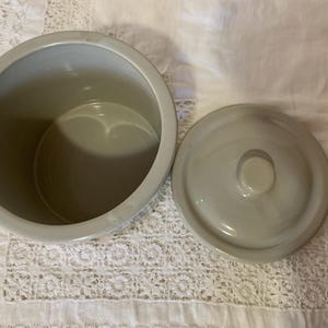 May include: A light gray ceramic jar with a matching lid. The jar is round with a wide opening, and the lid has a small knob. The jar and lid are on a white surface.