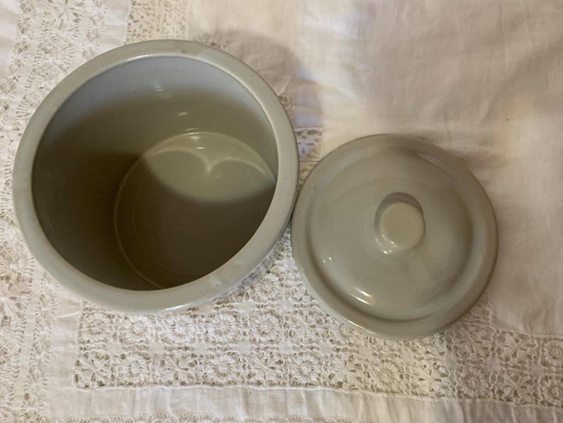 May include: A light gray ceramic jar with a matching lid. The jar is round with a wide opening, and the lid has a small knob. The jar and lid are on a white surface.