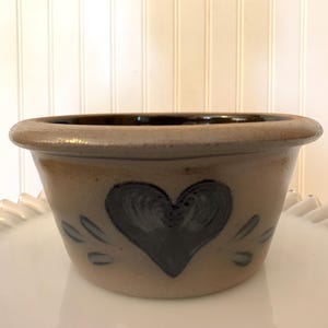 May include: A tan-colored ceramic bowl with a dark brown rim and a black heart design. The bowl has a rustic, handcrafted appearance, suitable for kitchen or home decor.