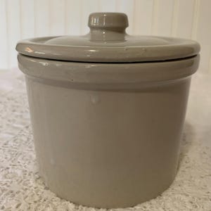 May include: A cylindrical ceramic crock with a smooth, light beige glossy finish. The container features a matching fitted lid with a central knob handle. The exterior surface is uniform in color with a subtle sheen, showing a simple, functional design.