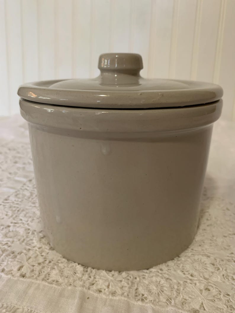 May include: A cylindrical ceramic crock with a smooth, light beige glossy finish. The container features a matching fitted lid with a central knob handle. The exterior surface is uniform in color with a subtle sheen, showing a simple, functional design.