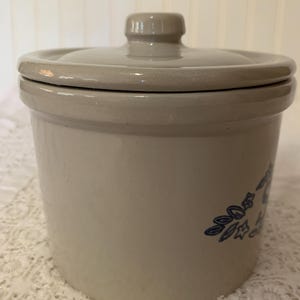 May include: A beige ceramic crock with a matching lid. The crock features a blue floral design on the side. The lid has a small knob for easy handling. The crock is likely used for food storage.
