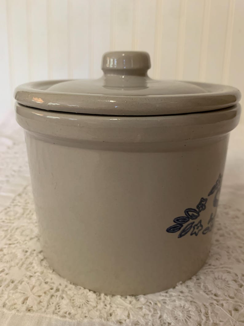 May include: A beige ceramic crock with a matching lid. The crock features a blue floral design on the side. The lid has a small knob for easy handling. The crock is likely used for food storage.
