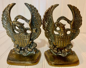May include: Pair of vintage brass bookends featuring an eagle with outstretched wings, a shield, and the inscription "E Pluribus Unum." The bookends are rectangular and have a weathered, antique appearance. Each bookend measures approximately 5 inches tall.