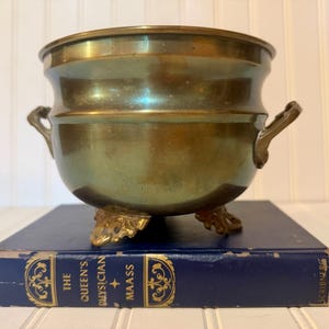 May include: A polished brass bowl with a rounded body, two side handles, and three ornate feet. The bowl features a smooth, metallic finish with subtle golden and greenish tones, resting on a dark blue book titled The Queen's Physician by Maass.