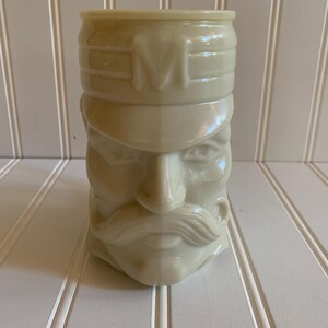 May include: A white ceramic mug shaped like a man's face with a mustache and a hat. The hat has the letter "M" on the front.