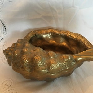 May include: A gold-colored brass seashell ashtray with a detailed, textured surface. The shell is open and has a wide, shallow bowl.