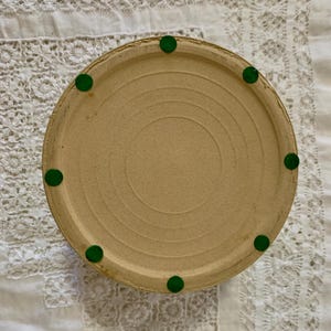 May include: A round, beige ceramic piece with a series of concentric circles on the bottom. The piece has a slightly raised edge and is supported by eight small, round, green feet. The piece is resting on a white, lace-trimmed surface.
