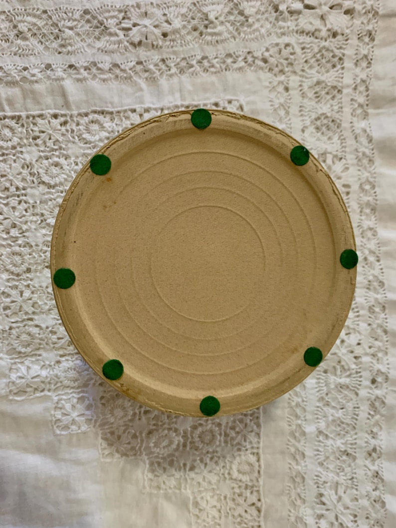 May include: A round, beige ceramic piece with a series of concentric circles on the bottom. The piece has a slightly raised edge and is supported by eight small, round, green feet. The piece is resting on a white, lace-trimmed surface.