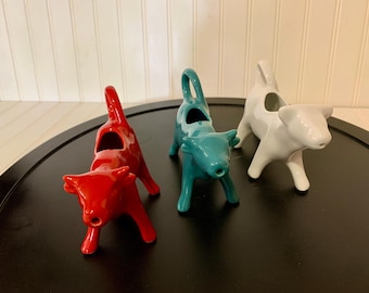May include: Three ceramic cow creamers in red, teal, and white. Each creamer has a handle and a spout, and is shaped like a cow. The creamers are on a black surface against a white background.